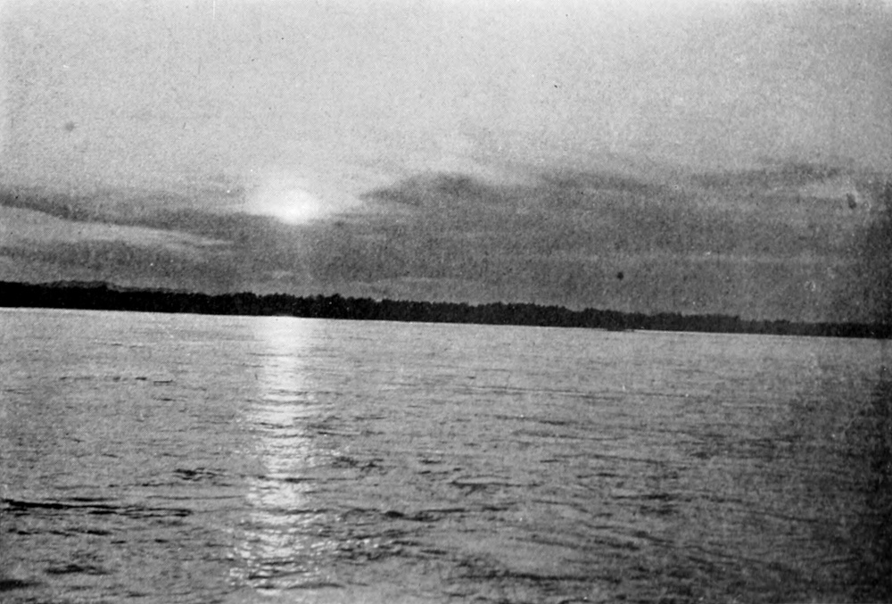 Sunset, with reflection of sun across a wide river; a dark shoreline is in the distance.
