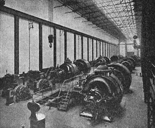 A large power station turbine hall — a long industrial interior with rows of massive generators or alternators on both sides under a glass roof — typical of early 20th-century electricity generating stations.