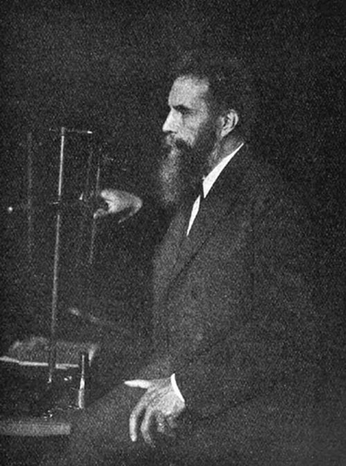 A photograph of a bearded man in profile seated beside scientific apparatus — consistent with portraits of Wilhelm Röntgen, discoverer of X-rays in 1895.