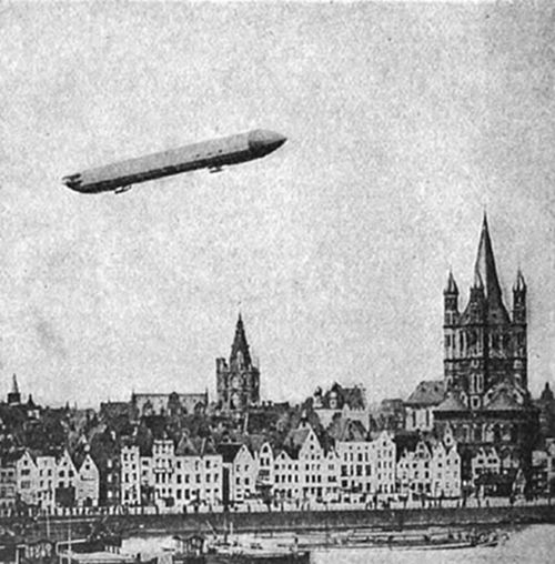 A photograph of a Zeppelin airship flying over a German city skyline with Gothic church spires and gabled rooftops — likely Cologne — an early 20th-century image of rigid airship travel over the Rhine.
